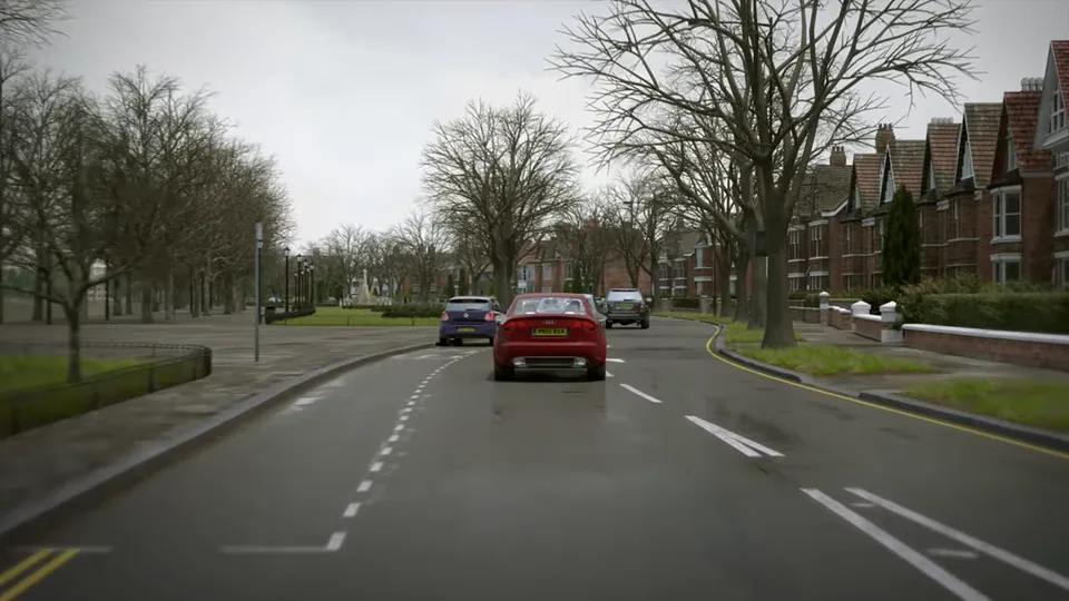 A wide avenue in winter runs between a lamp-lined park and red-brick terraced houses, with light traffic ahead and bare street trees.