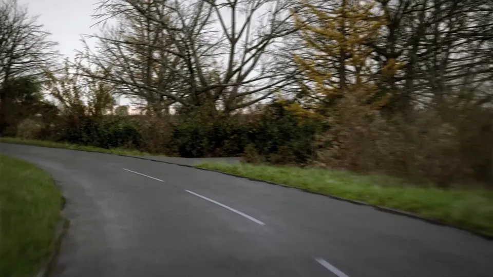 A narrow rural lane bounded by tall hedgerows and fenced fields, crossing a low stone-parapet bridge under a grey sky.