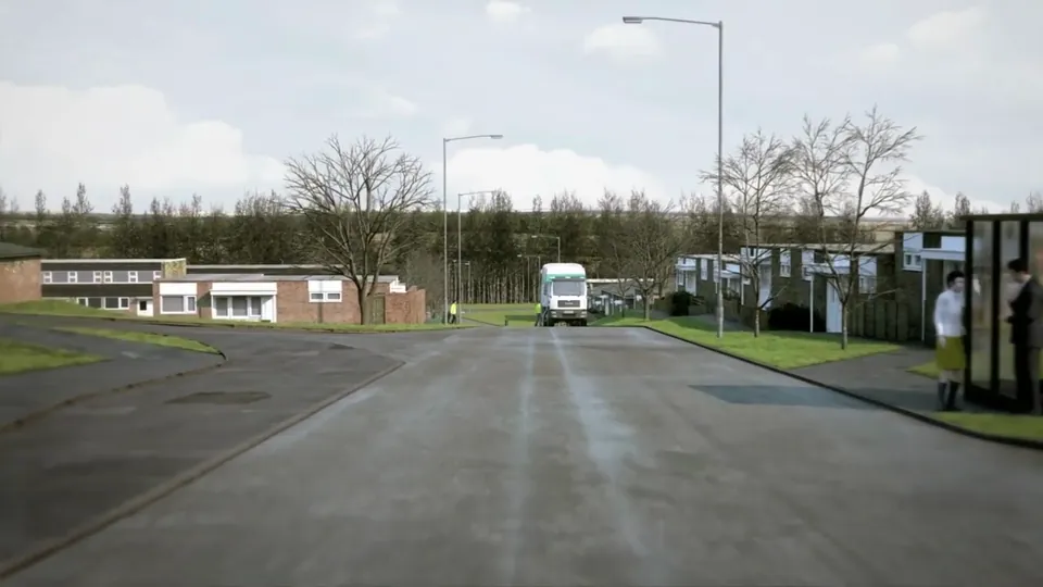 A wide low-rise estate road flanked by single-storey brick buildings, broad grass verges and avenues of bare poplar trees.