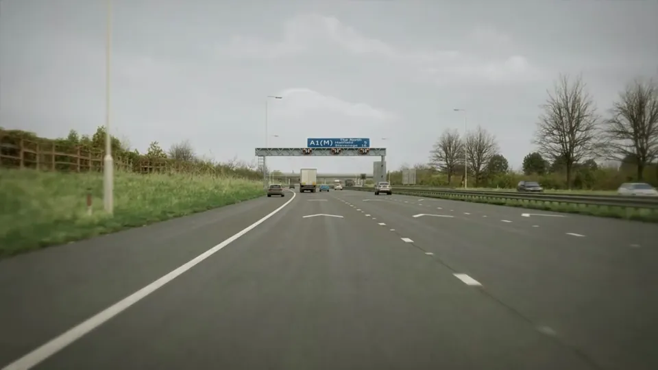 A multi-lane motorway with overhead direction gantries, grass verges and hatched road markings under a grey sky.