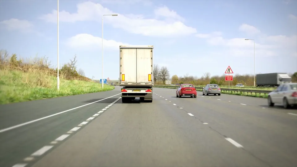 A multi-lane motorway with a central barrier and grass verges runs beneath overhead road bridges under a clear blue sky.