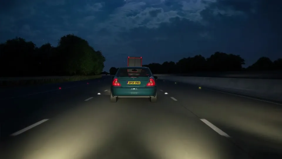 A multi-lane motorway at night with reflective cat's-eye studs and overhead gantry signs, bordered by dark tree-lined verges.