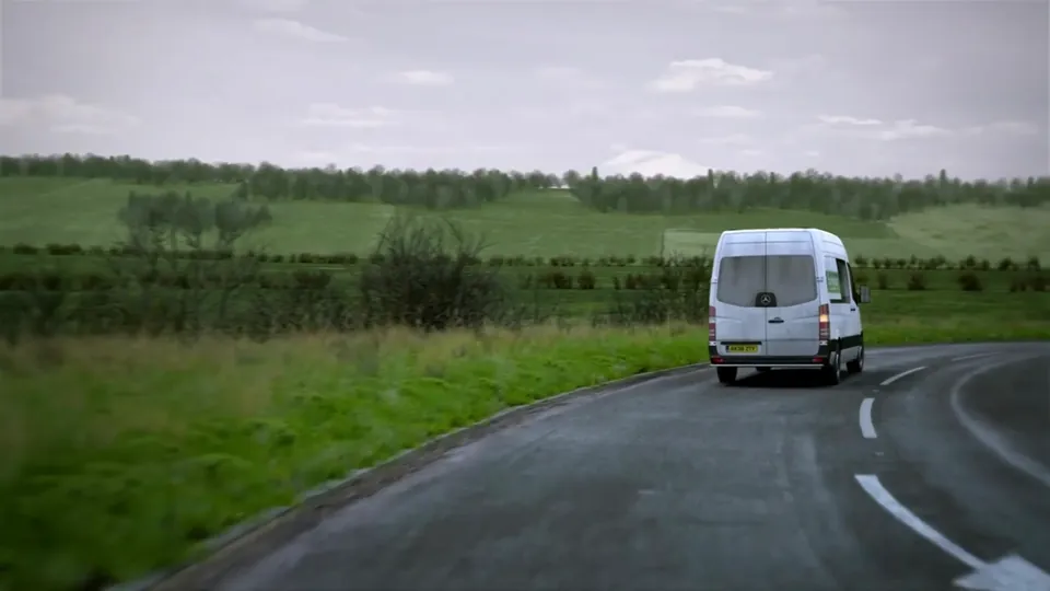 An open rural A-road sweeps through rolling green fields and hedgerows with scattered bare trees on a damp day.
