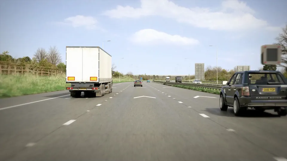 A wide motorway with overhead gantry direction signs, grass verges and hedgerows under a bright blue sky.
