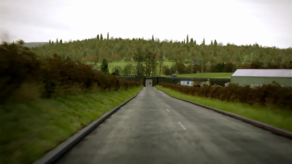 A country road dips beneath a narrow single-track railway underpass and crosses a stone hump-back bridge through open parkland.