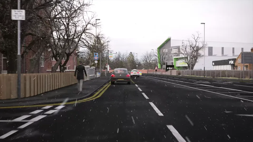 Snow falls over a busy town road beside a timber fence and red-brick houses, with queueing cars, roadside signs and retail units ahead.