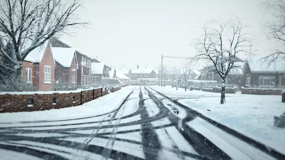 A residential street blanketed in snow during heavy snowfall, with snow-covered houses, garden walls and bare trees, tyre tracks worn through the snow.