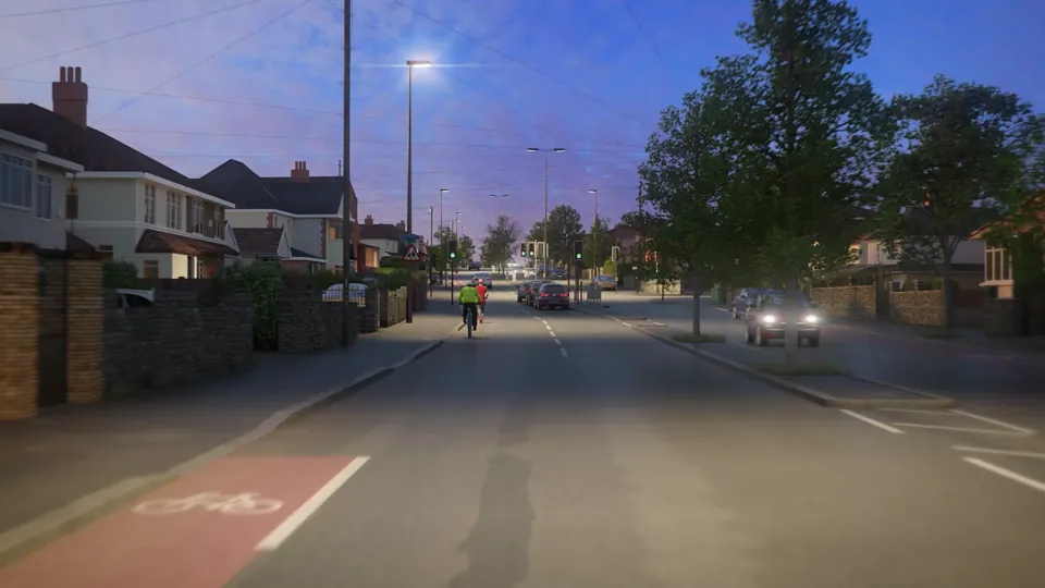A wide suburban through-road at twilight, lined with houses, lit street lamps, red cycle lanes and a traffic-light junction under a purple sky.