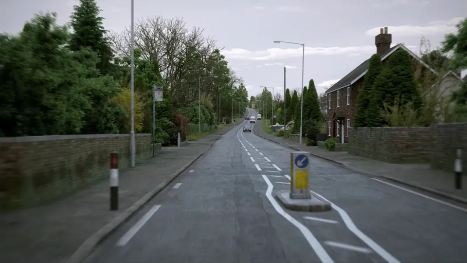 A wide edge-of-town residential road with hatched centre markings and a Belisha-beacon zebra crossing, bordered by semi-detached houses.