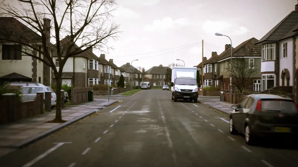A suburban residential street of semi-detached houses with both kerbs lined by parked vehicles and bare winter trees.