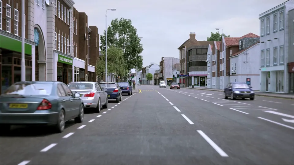 A wide town high street lined with two-storey shopfronts, parked cars, traffic lights and street trees.