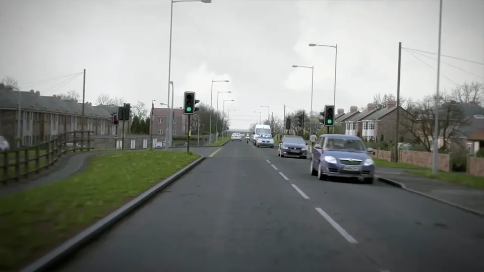 An urban road lined with brick houses and bungalows, street lighting, traffic lights and yellow box-junction markings.