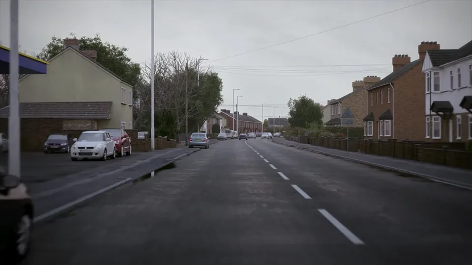 A wide town road passes red-brick semi-detached houses, with parked cars in a lay-by, a filling-station canopy and traffic in the far distance.