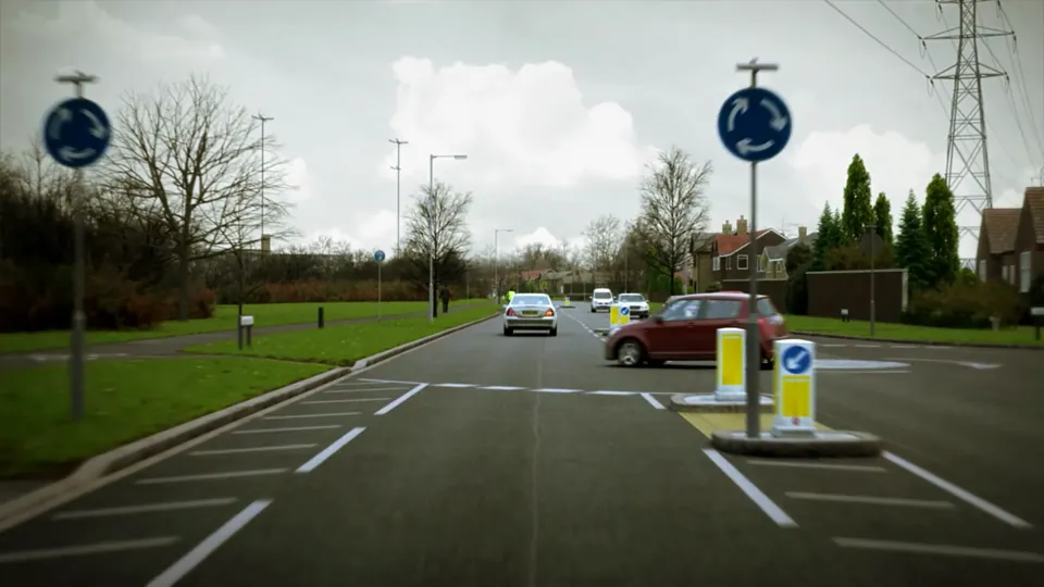 A wide urban distributor road with central hatched markings, pedestrian refuge islands and bollards, houses set back behind verges.