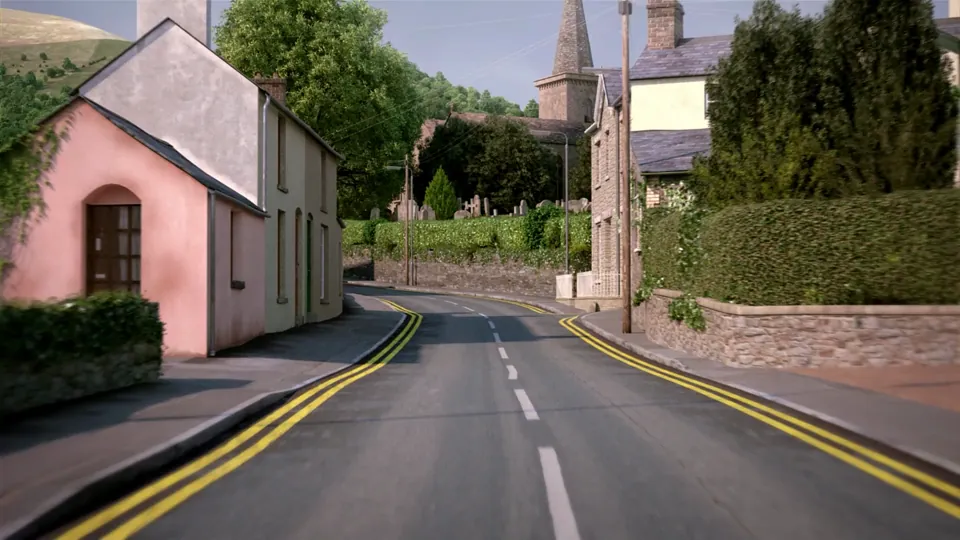 A curving downhill village street set against a steep green hillside, with stone walls, whitewashed cottages and a church spire.