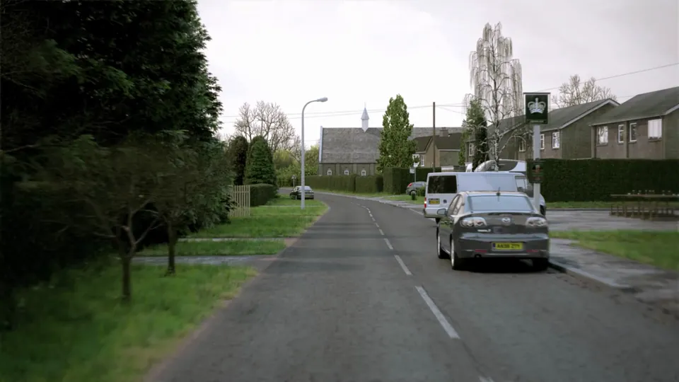 A village-edge road passes detached houses with neat clipped hedges and front gardens beneath a spired church.