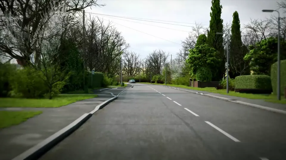A road leaves a hedged village and opens out toward bare-treed countryside with grass verges and roadside daffodils.