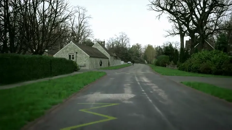 A narrow village lane lined with stone cottages, a war-memorial cross and clipped hedges, the road wet with puddles.