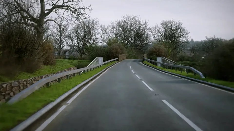 A damp winter road rises over a small bridge flanked by metal crash barriers and white parapets, with a dry-stone wall and dense bare trees along the verge.
